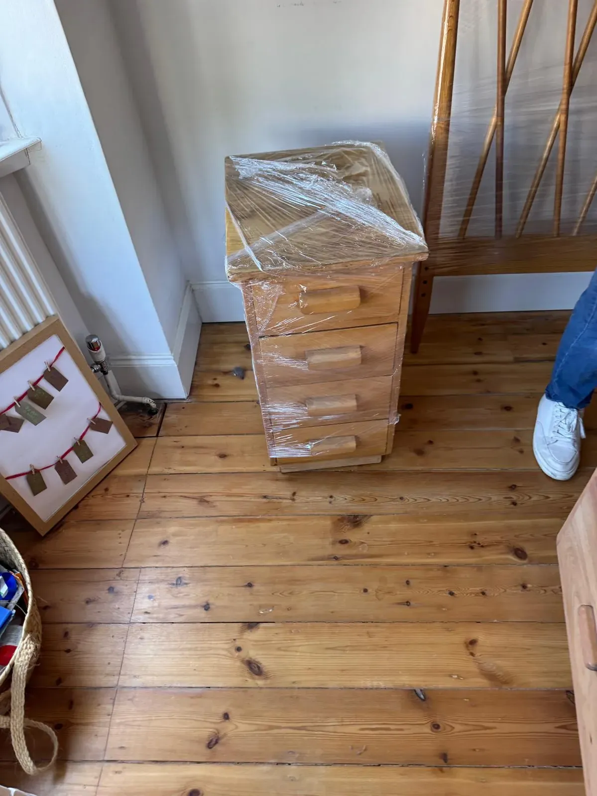 A pine bedside chest of drawers wrapped in cling film on bare floorboards, next to a wrapped wooden headboard