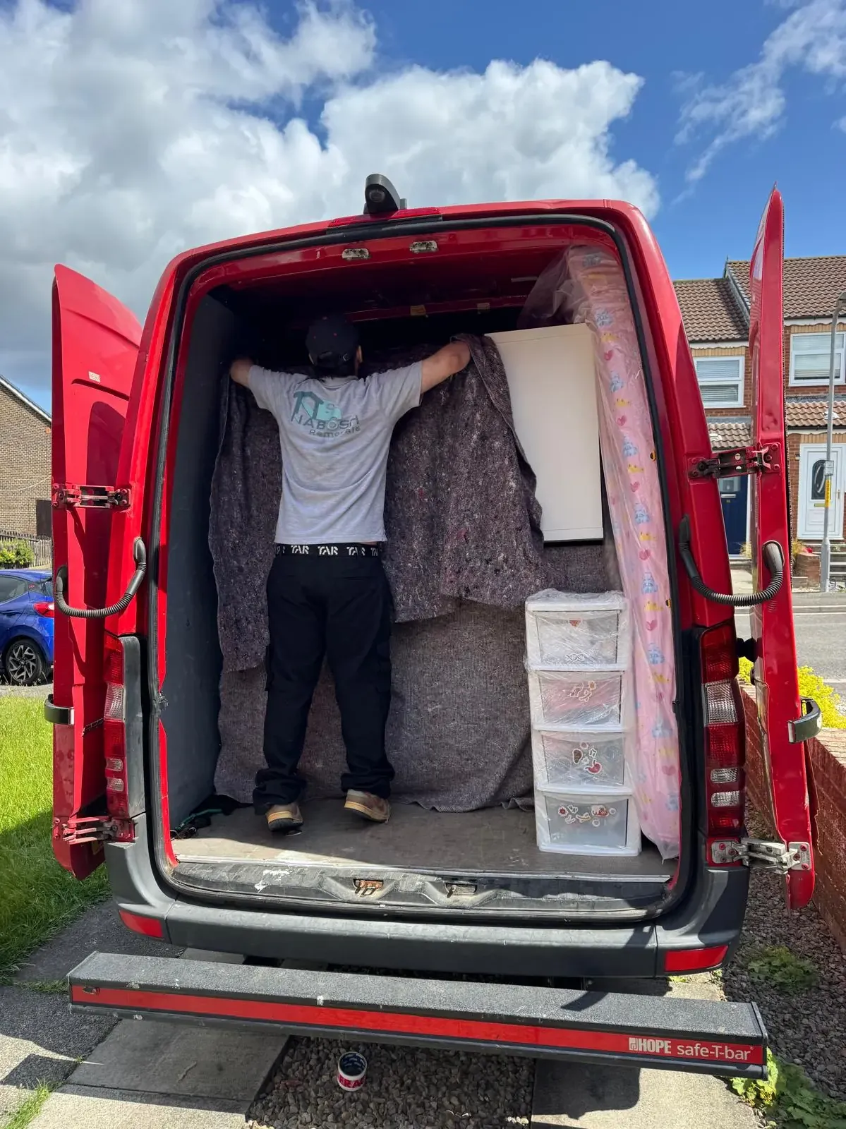 A crew member in a Nabosh Removals T-shirt standing in the back of a red van, pulling a moving blanket over a tall piece of furniture, with a plastic drawer unit and a wrapped mattress beside him