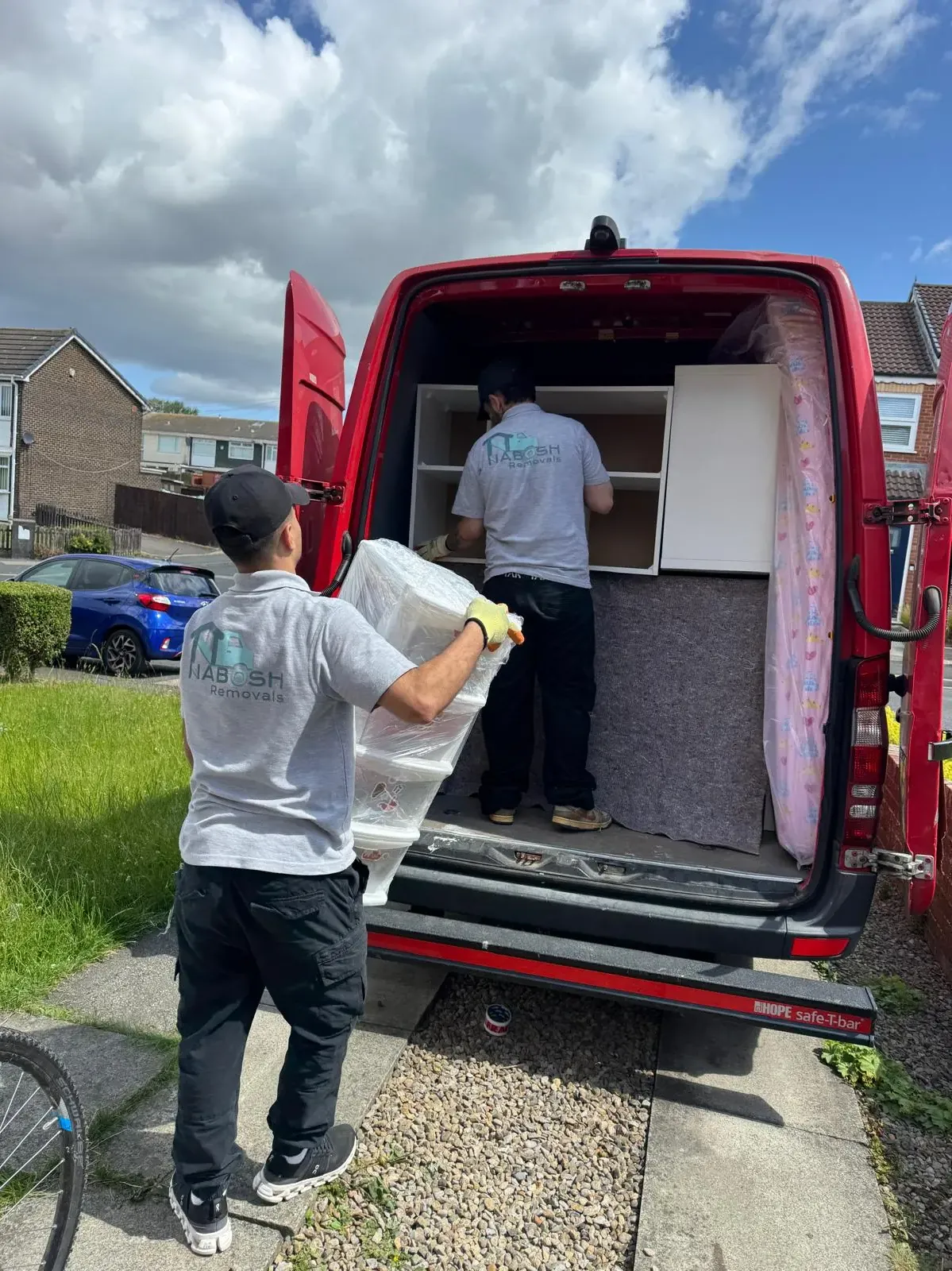 A crew member carrying cling-filmed plastic drawer units to a red van while a second crew member stacks white shelving units inside
