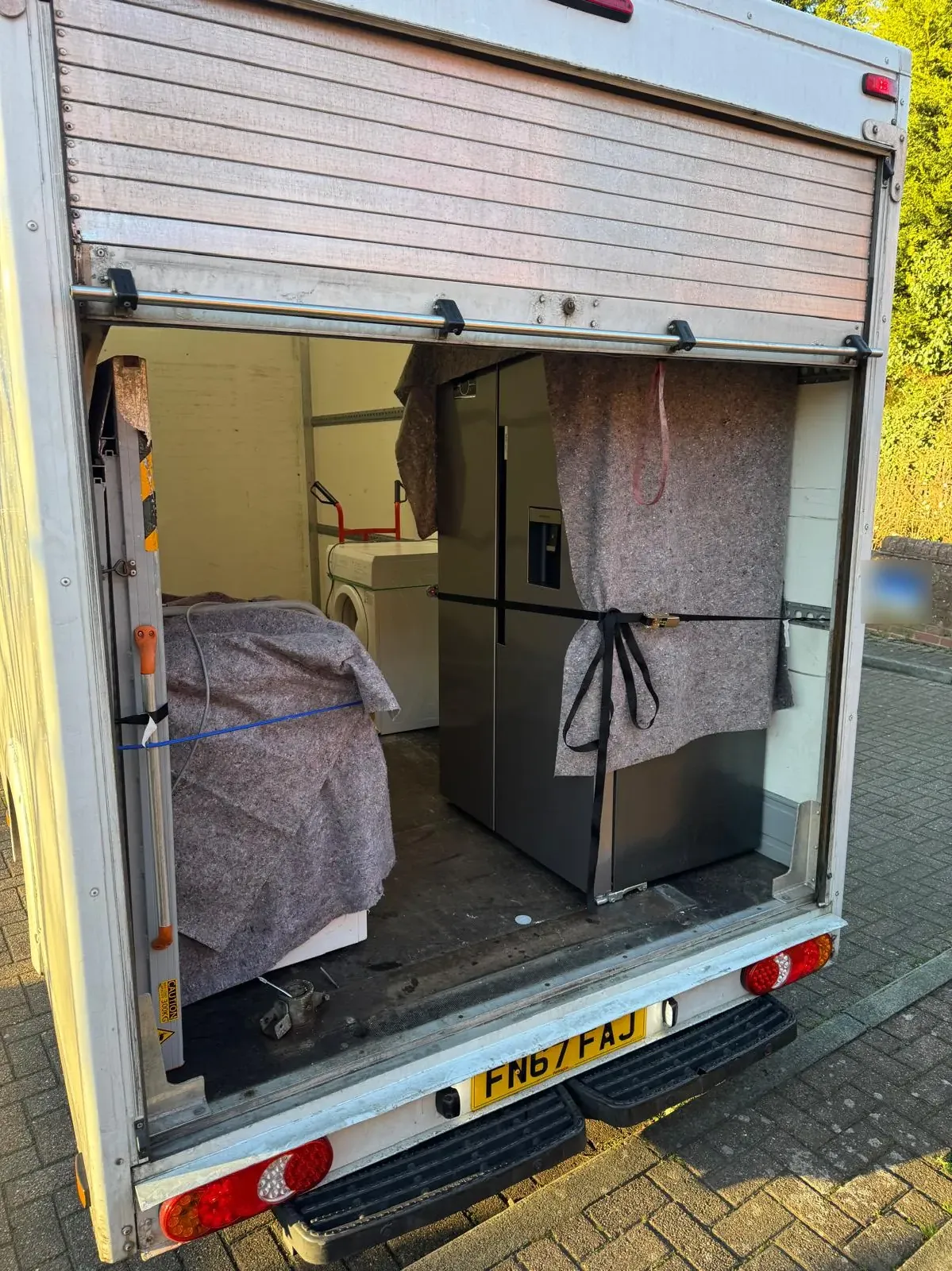 An American-style fridge freezer under a moving blanket, ratchet-strapped upright to the side of a Luton van, with a washing machine and blanket-covered appliances loaded beside it
