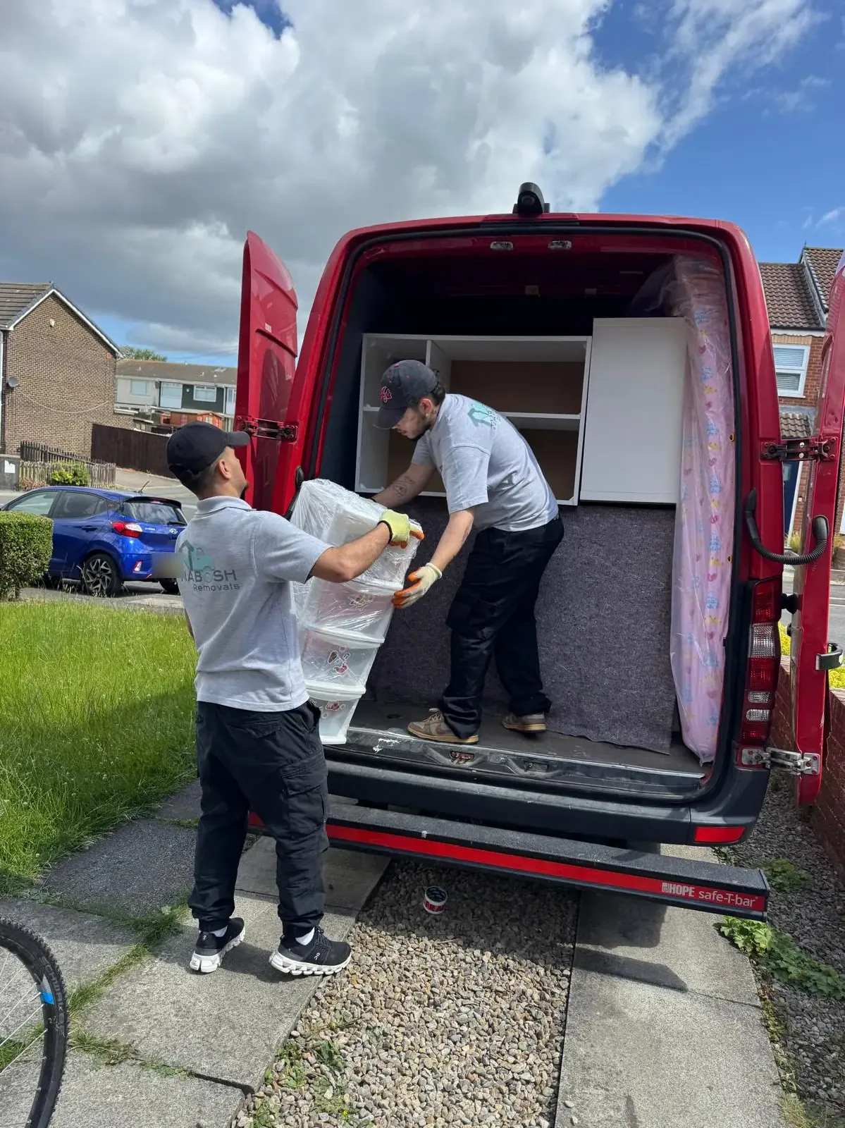 Two crew members in Nabosh Removals T-shirts and work gloves passing cling-filmed plastic drawer units into the back of a red van parked on a driveway