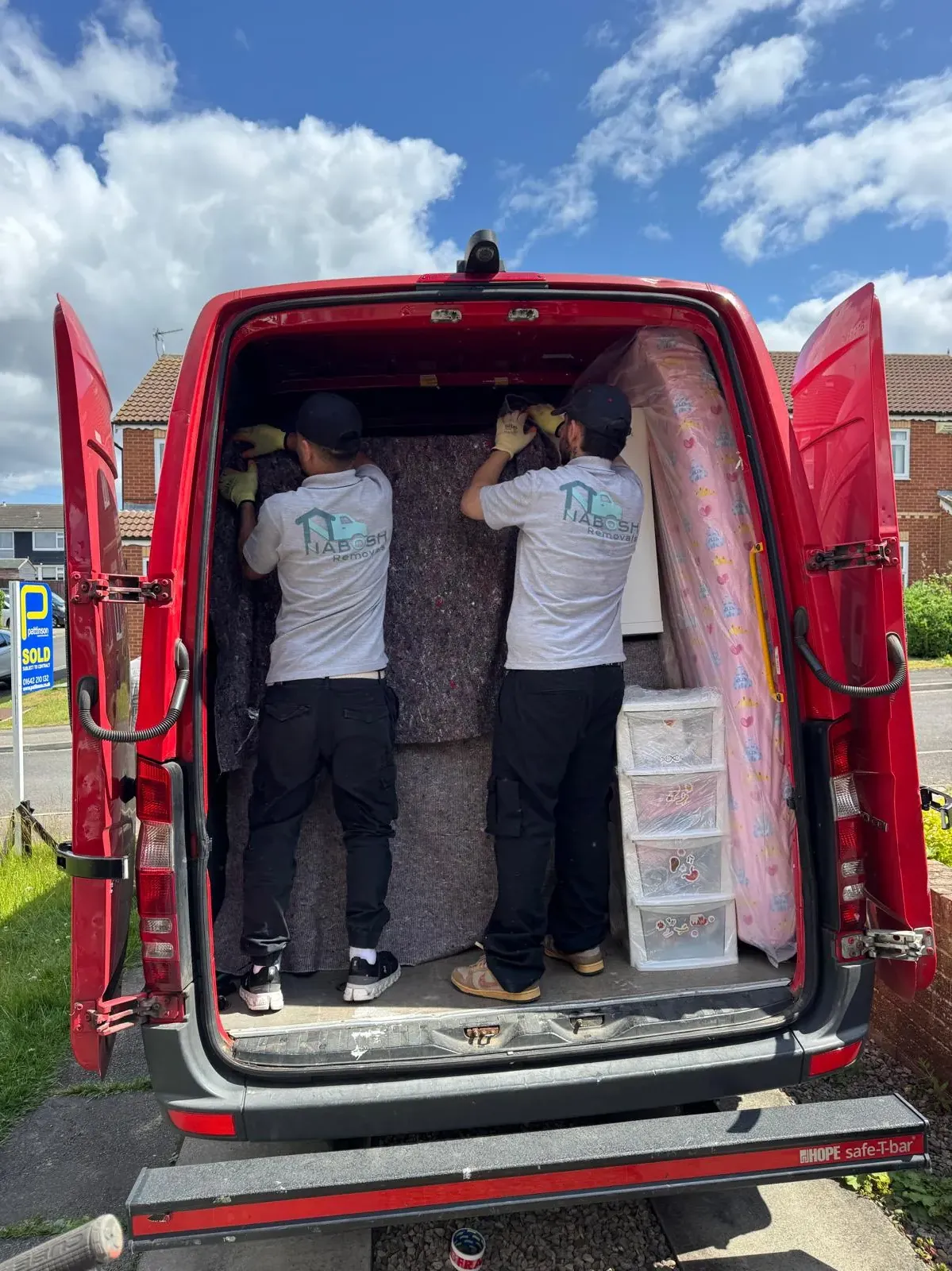 Two crew members in Nabosh Removals T-shirts and work gloves hanging moving blankets inside a red van, beside a wrapped mattress and a plastic drawer unit