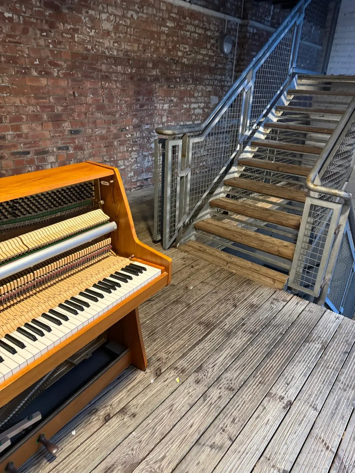 An upright piano with its front panel off, showing the keys and hammers, standing on wooden decking beside an open metal staircase