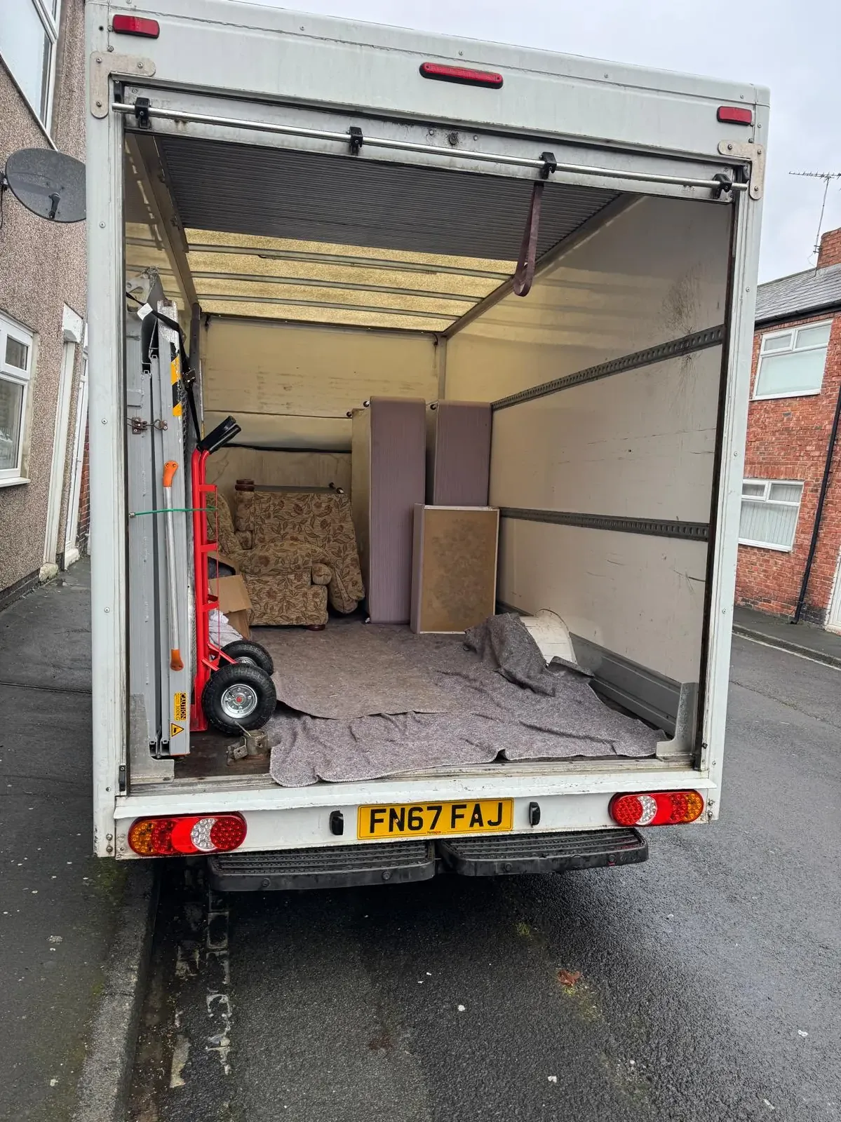 The back of a Luton van on a terraced street, with an armchair, mattresses and a headboard loaded at the front, moving blankets on the floor and a sack truck stowed by the tail lift