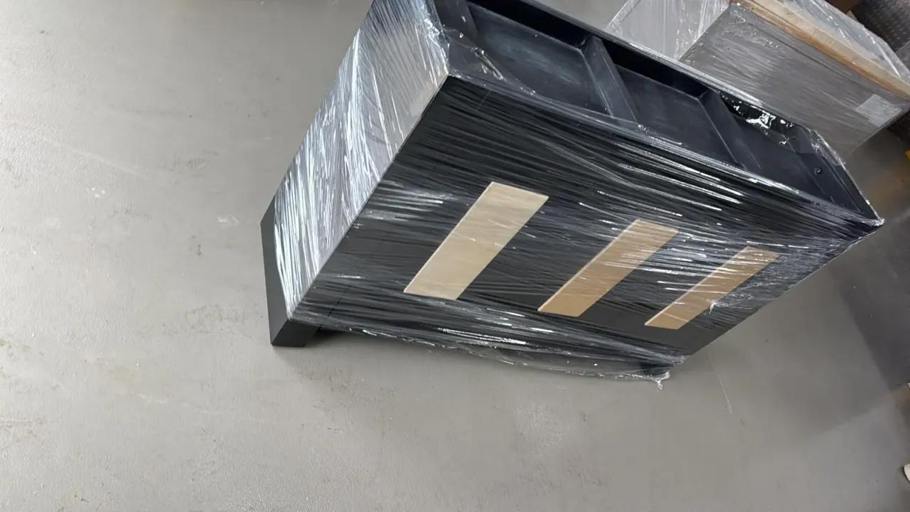 A black sideboard with pale wooden handles, wrapped in stretch film and standing on a concrete floor