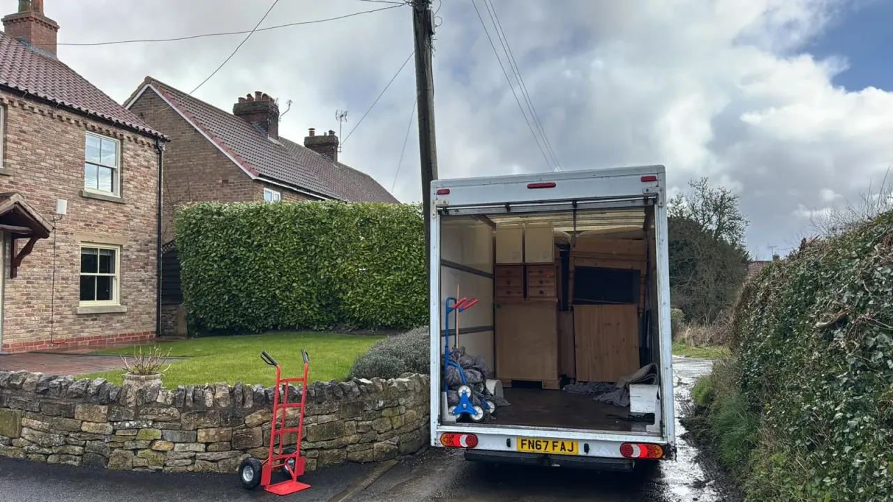 The back of a Luton van open on a country lane outside a brick house, loaded with pine wardrobes and chests of drawers, with a red sack truck beside a low stone wall