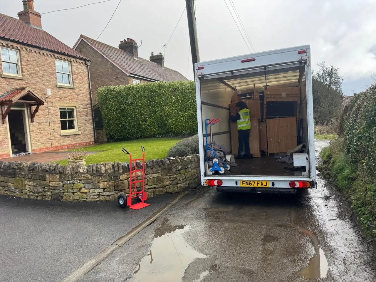 A Luton van parked on a wet village lane beside a low stone wall, with a crew member in a hi-vis vest loading pine furniture inside and a red sack truck on the road