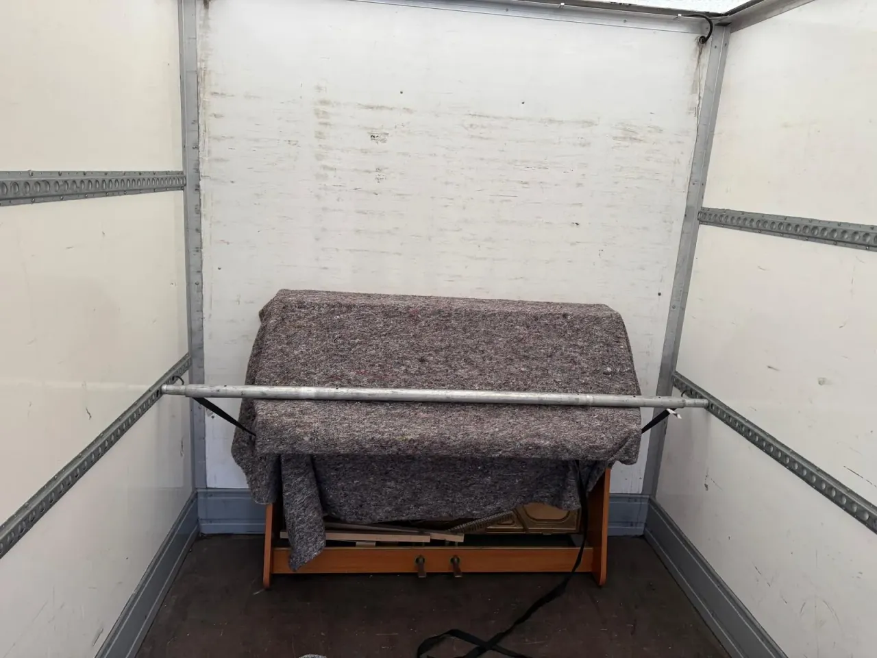 An upright piano under a moving blanket at the front of an otherwise empty van, held in place by a load bar and straps