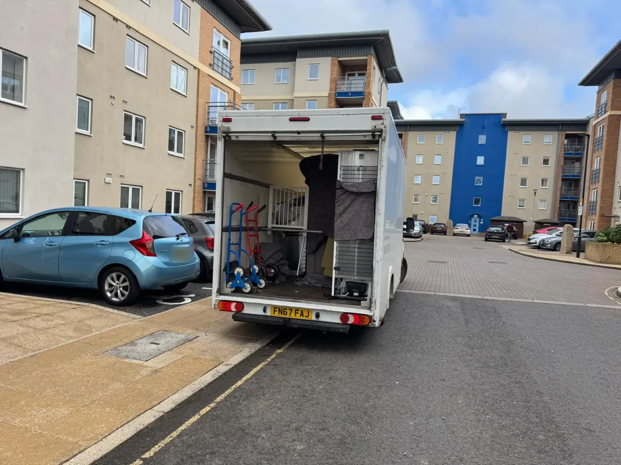 A Luton van parked outside a block of flats with its shutter up, carrying a blanketed fridge freezer, a white cot and sack trucks