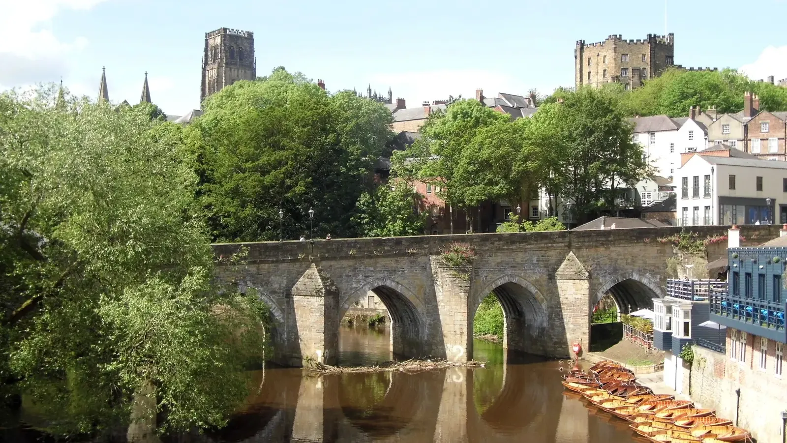 Elvet Bridge over the River Wear in Durham on a summer day, with rowing boats moored beside it, the cathedral tower above the trees on one side and the castle keep on the other
