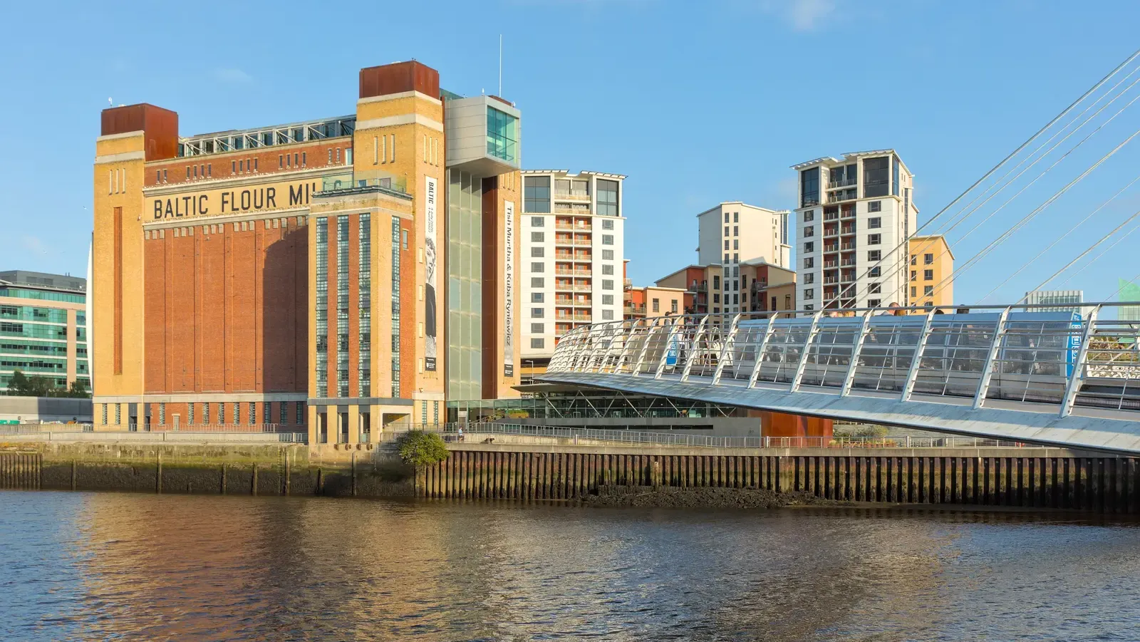 The Baltic Centre for Contemporary Art on the Gateshead bank of the Tyne in evening sun, with BALTIC FLOUR MILLS painted across its brick tower and the Millennium Bridge curving in beside it