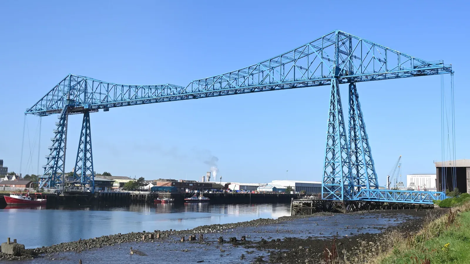The blue steel Transporter Bridge crossing the River Tees in Middlesbrough under a clear sky, seen from the Middlesbrough bank at low tide with fishing boats moored below it
