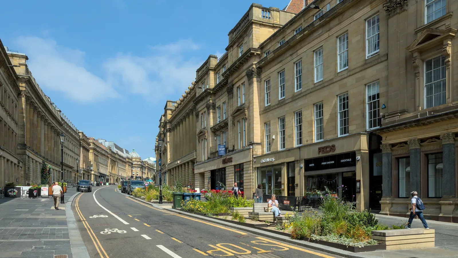 Grey Street in Newcastle on a sunny day, its curving terrace of classical stone buildings running down towards the city centre, with planted beds and a cycle lane in the foreground